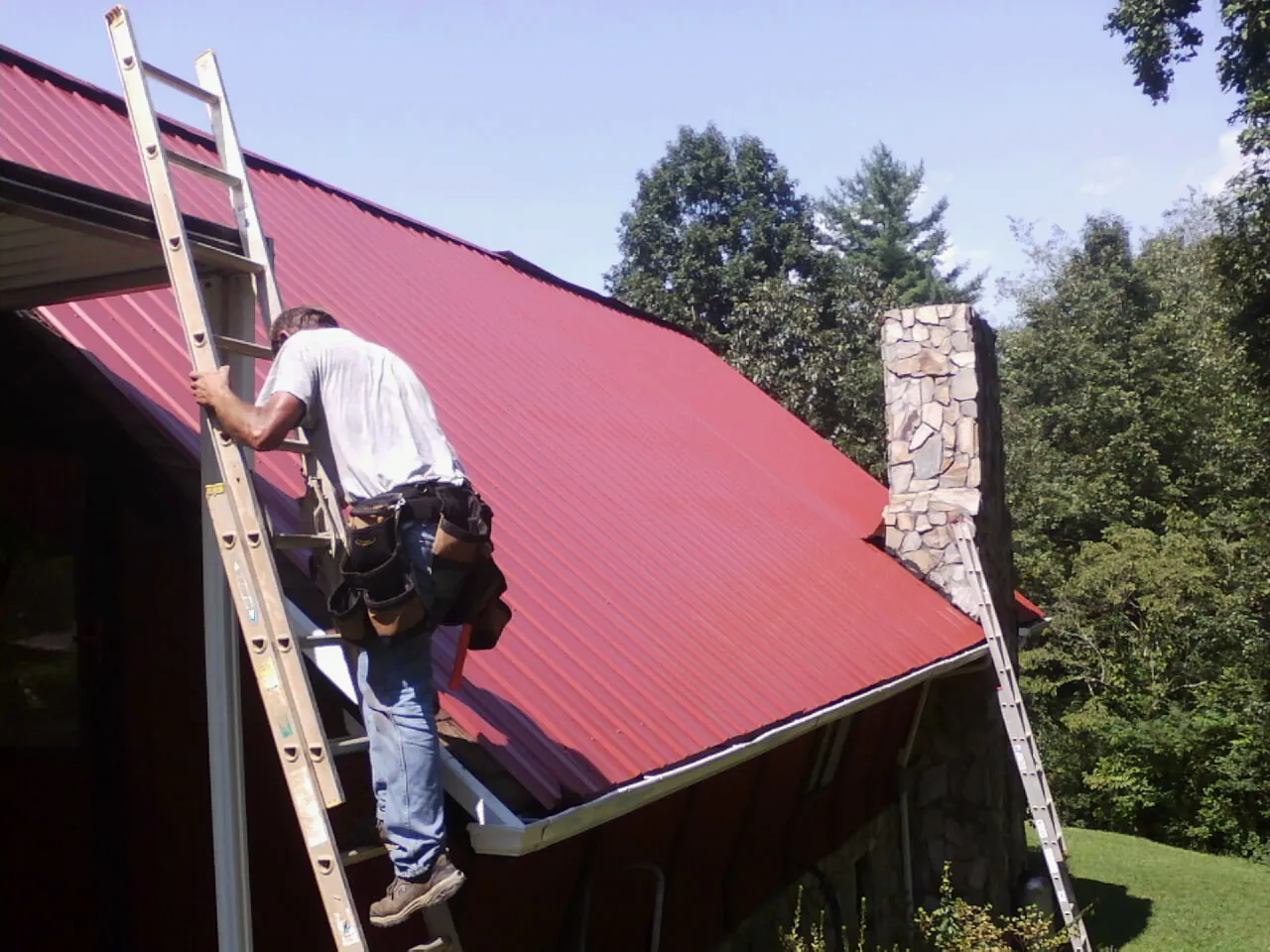 Roofer on a ladder beside a red metal roof and stone chimney — Soffit & Fascia Repair by Roofing Brothers in Woodacre